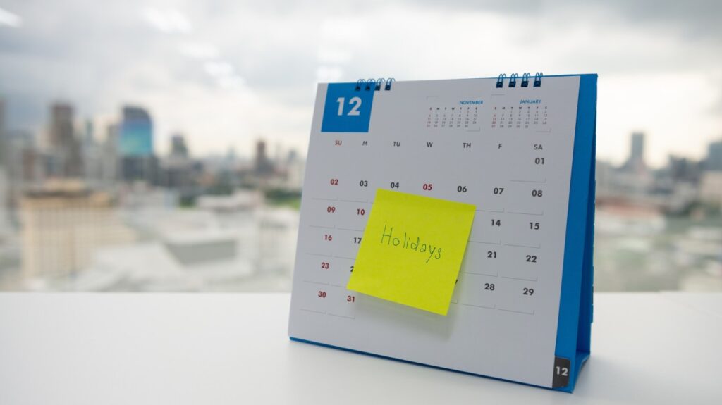 A desk calendar displaying the month of December with a yellow sticky note that reads "Holidays" attached to it. The calendar is placed on a white surface with a blurred cityscape visible in the background.